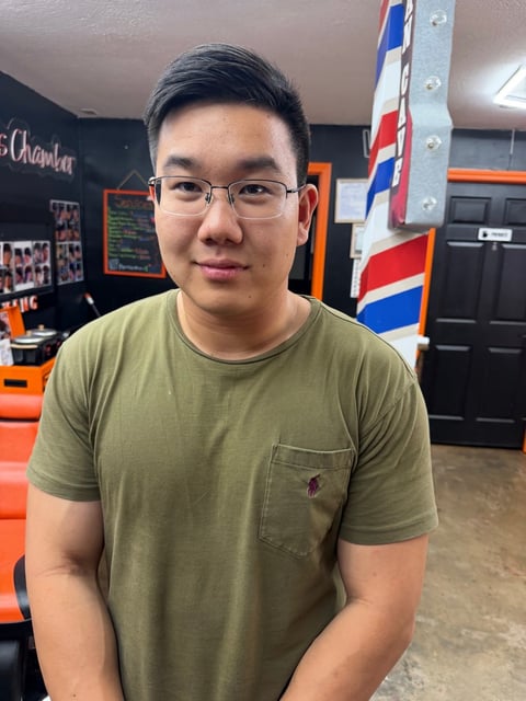 Man wearing glasses and olive green t-shirt standing indoors in front of orange furniture and Thai flag