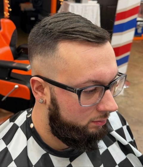 Man with styled undercut haircut, dark beard, and glasses wearing a checkered shirt in a barber shop setting