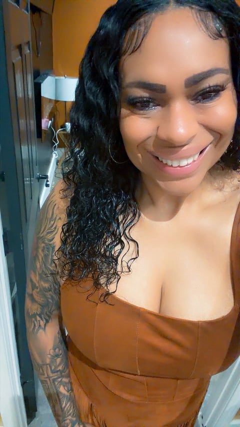 Smiling woman with long wavy black hair, tattoos on arm, wearing rust-colored top in indoor setting