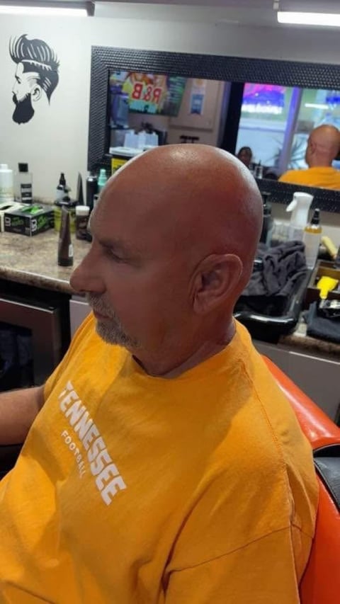 Bald man with goatee wearing yellow shirt sitting in barber chair, with mirror and barbershop visible in background