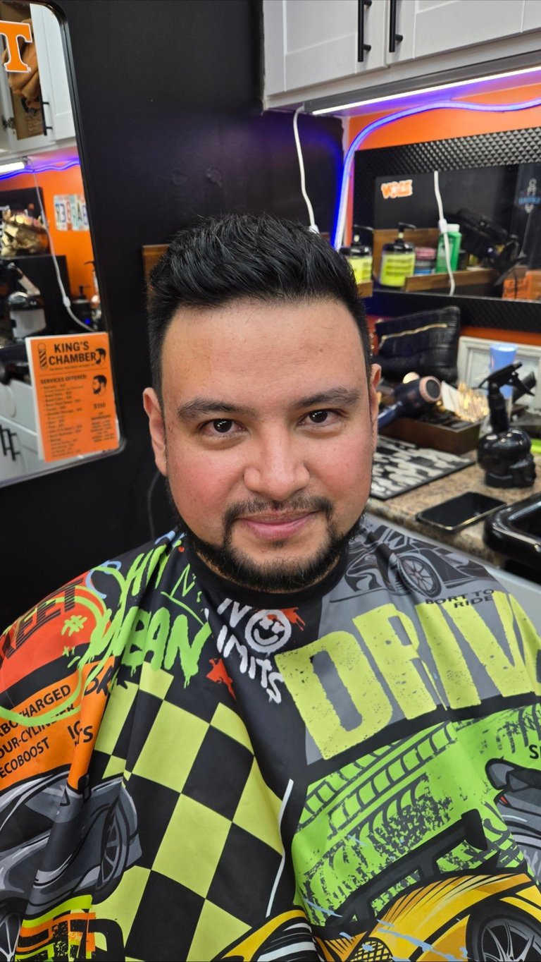 Man with slicked-back black hair wearing neon green and black graphic shirt, smiling at camera in gaming or retail store environment