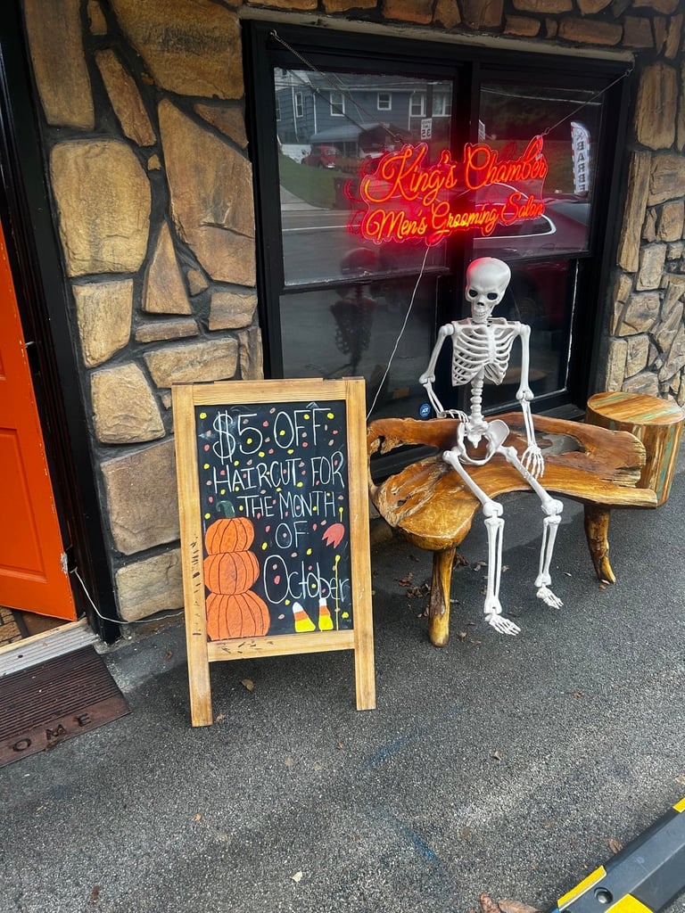 Halloween storefront display with skeleton sitting on wooden chair, red neon sign, and chalkboard sign advertising haircut deals