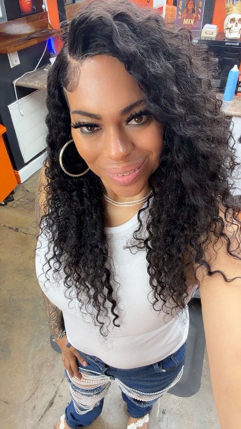Portrait of a woman with long curly black hair, gold hoop earrings, wearing a white tank top and distressed blue jeans, smiling at the camera